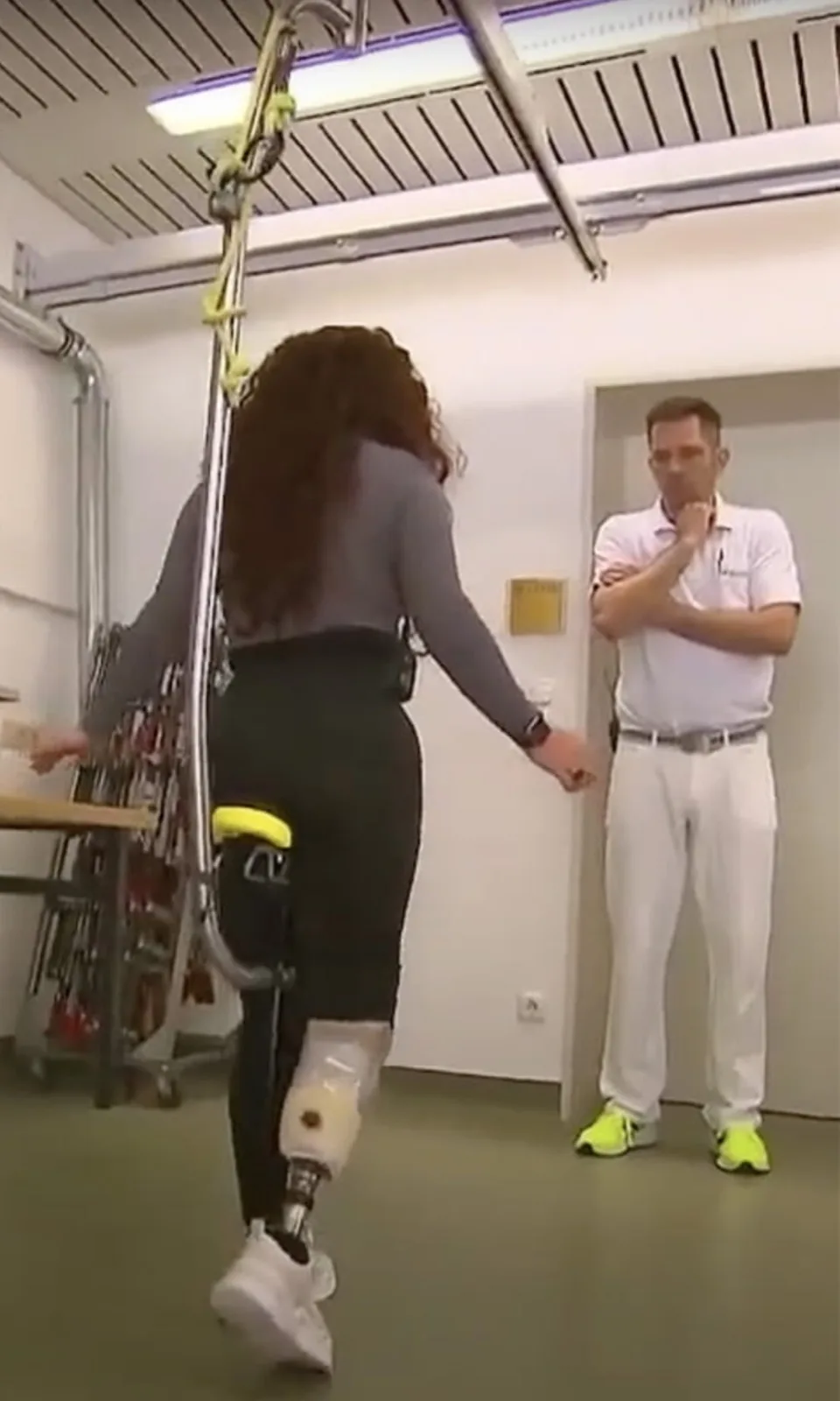 Vrouw met een beenprothese oefent het staan in de mobiliteitsbeugel, met een behandelaar ernaast
